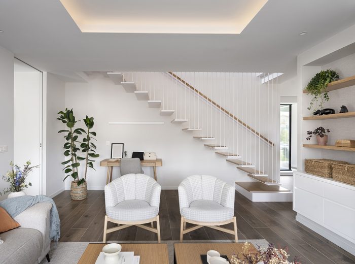 salón amueblado con sofá butacas mesa de café en tonos neutros y terracota, mueble con estanterías empotradas en madera y escalera con barandilla atirantada en blanco, vivienda unifamiliar LAS, obra nueva y diseño de interiores y amueblamiento, Vitoria-Gasteiz