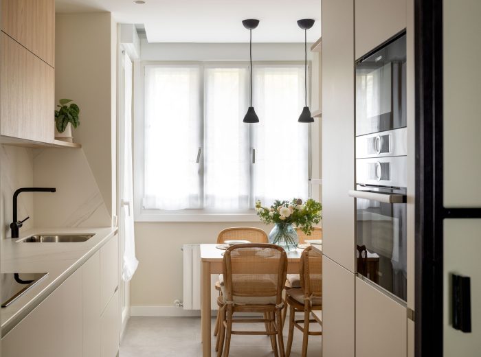 Cocina en color topo con electrodomésticos integrados y luminoso comedor al fondo, mesa de madera y sillas de ratán, reforma interior y diseño de interiores y amueblamiento EF13, Vitoria-Gasteiz