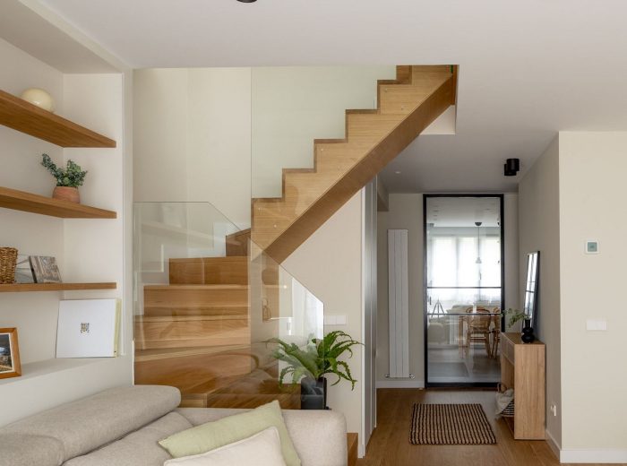 salón en tonos neutros y escalera escultórica en madera con cocina con cierre acristalado con perfilería de aluminio en negro al fondo, reforma interior y diseño de interiores y amueblamiento EF13, Vitoria-Gasteiz